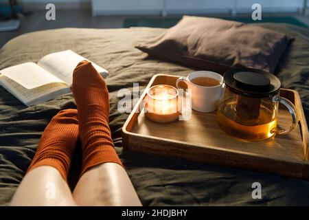 Soft photo of woman`s legs in the woolen socks on the bed with book and cup of tea and candle on the tray. Interior and home cosiness concept. Top vie Stock Photo